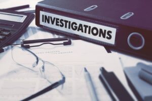 Ring Binder with inscription Investigations on Background of Working Table with Office Supplies, Glasses, Reports. 