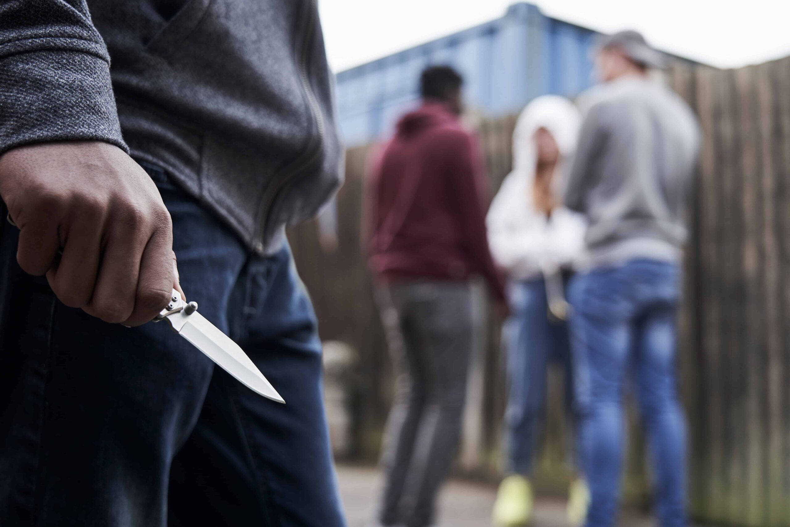 Person holding a knife in an alley with a group of people in the background, symbolizing assault or violent crime